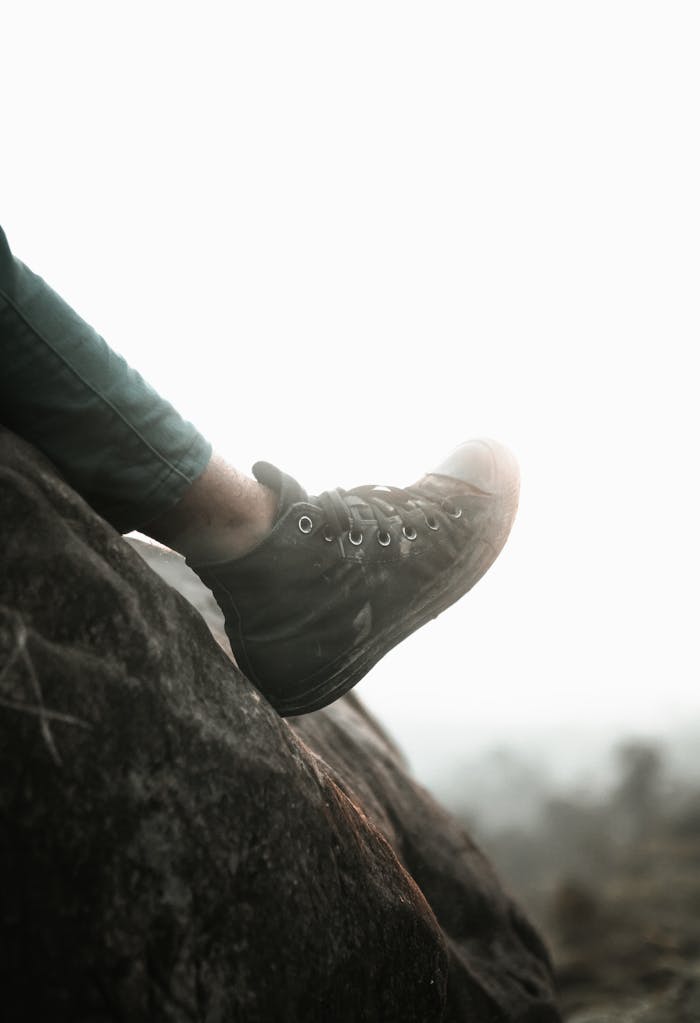 A person's foot wearing a sneaker resting on a rock cliff with a misty mountain view.